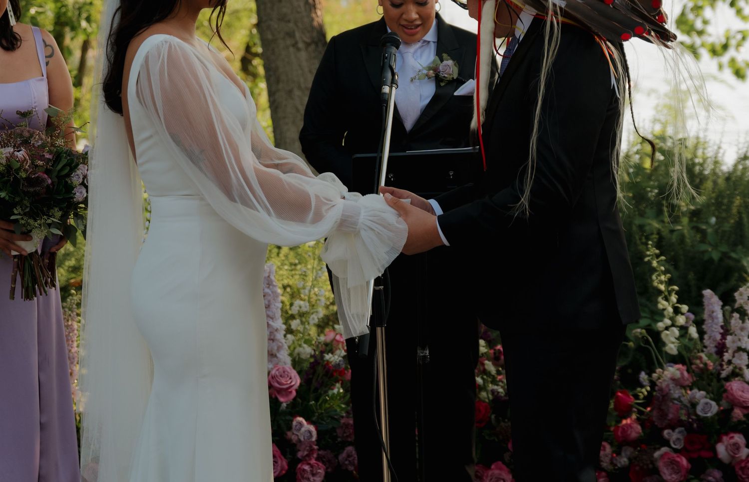 A couple exchanges wedding vows during an outdoor garden ceremony surrounded by blooming pink roses and greenery.