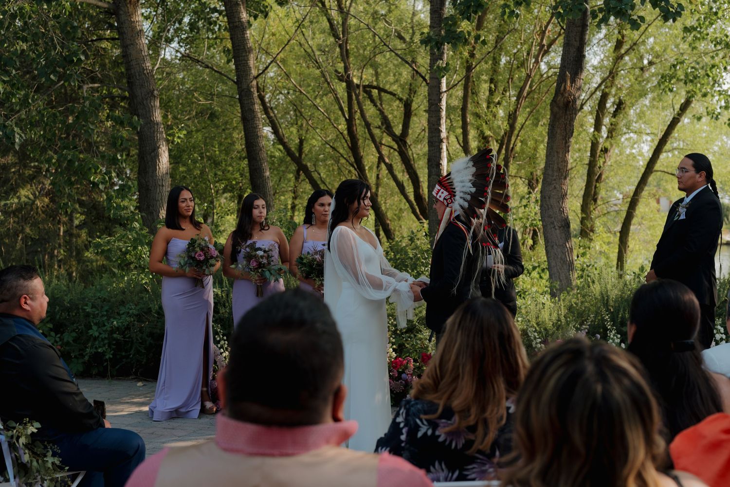 Outdoor wedding ceremony in a wooded area with guests seated on white chairs under green trees.