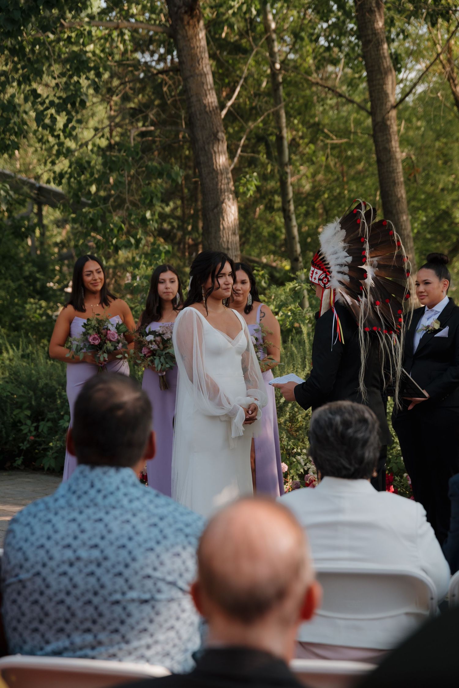 An outdoor wedding ceremony takes place in a wooded setting with attendees gathered to witness the occasion.