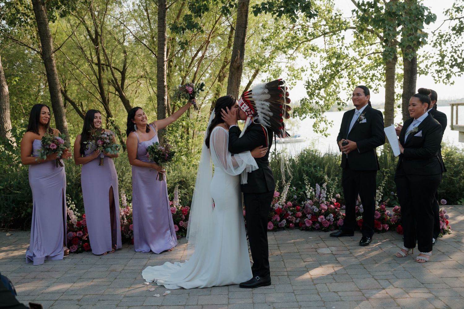 Wedding ceremony taking place outdoors under tall trees with guests seated on white chairs watching the proceedings.