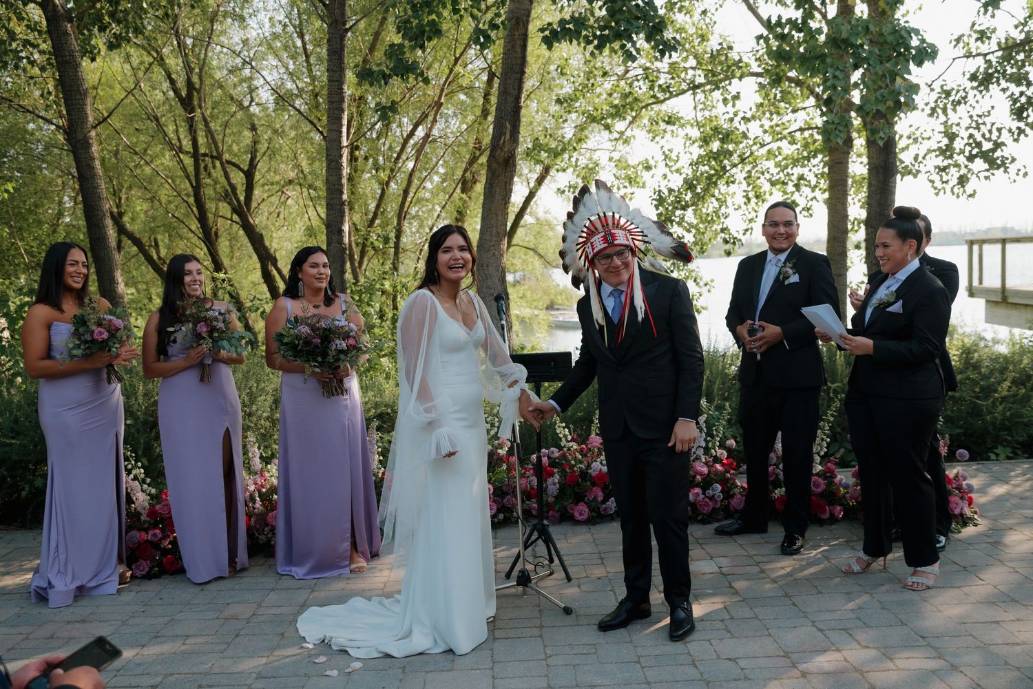 Wedding party in purple and black attire gathered outdoors under green trees for formal photographs.