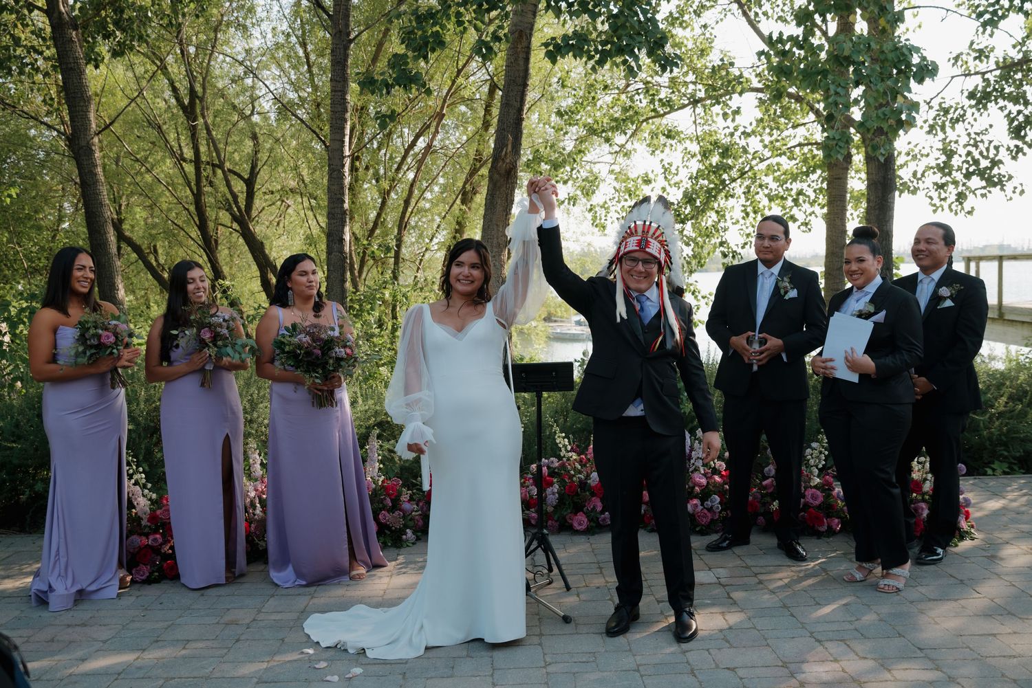 Wedding party stands in formation under shady trees during an outdoor ceremony.