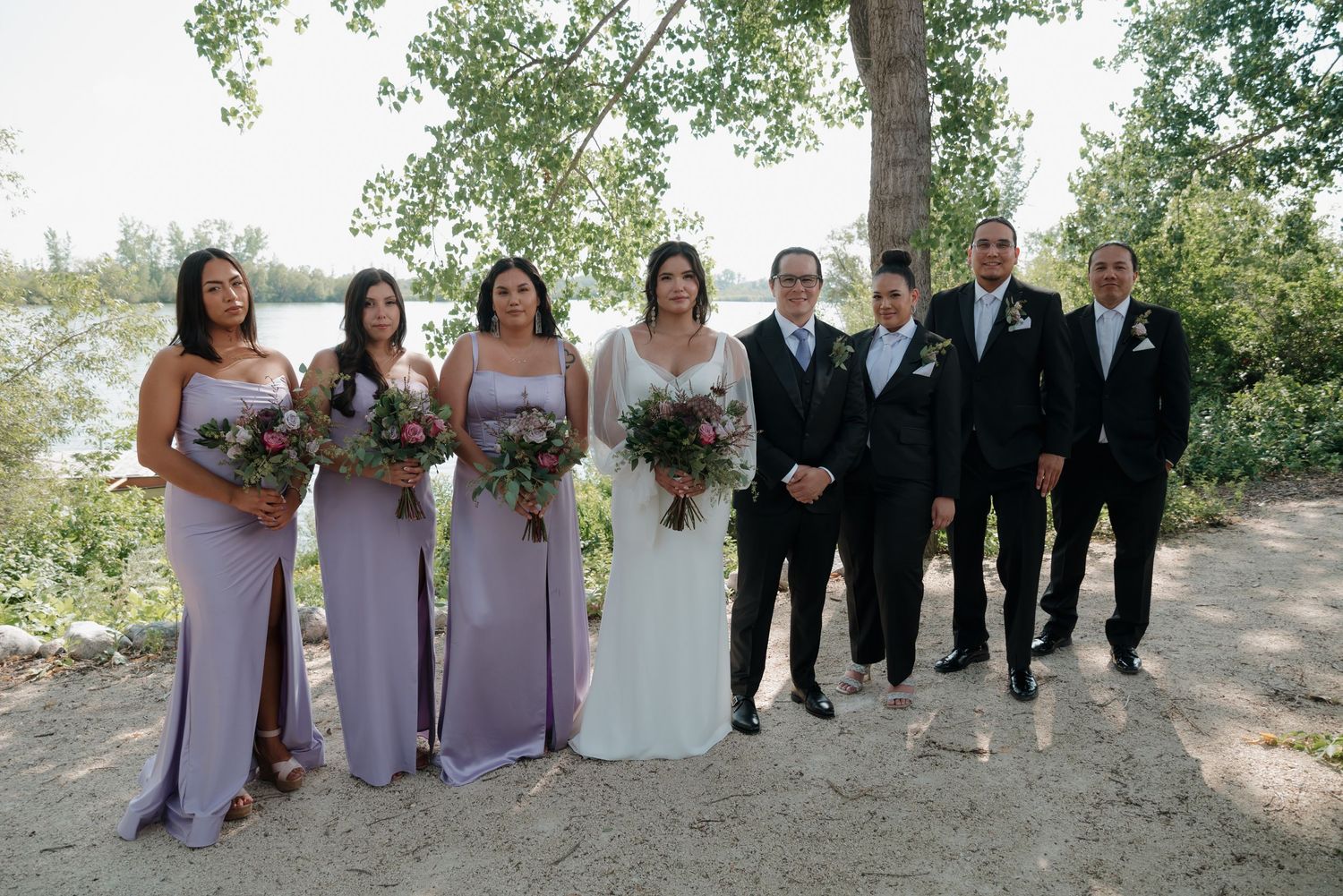 Wedding party in formal attire poses together on a dirt path surrounded by green trees and natural scenery.