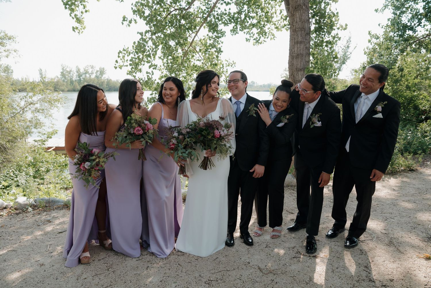 Wedding party in purple bridesmaid dresses and black suits pose together on a garden path.