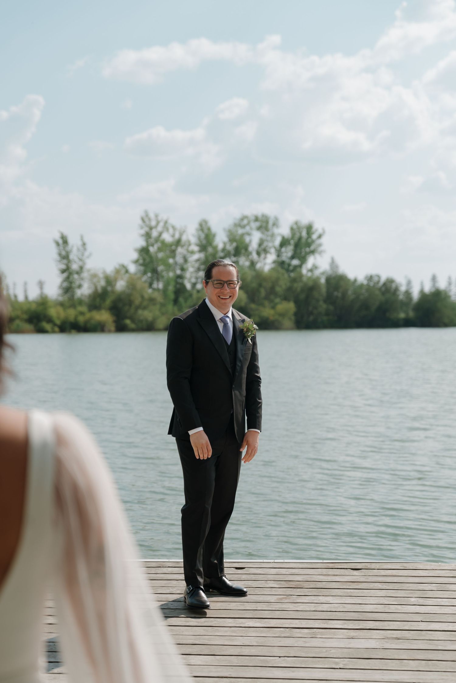 A person in a black suit stands on a wooden dock overlooking a serene lake with trees in the background.