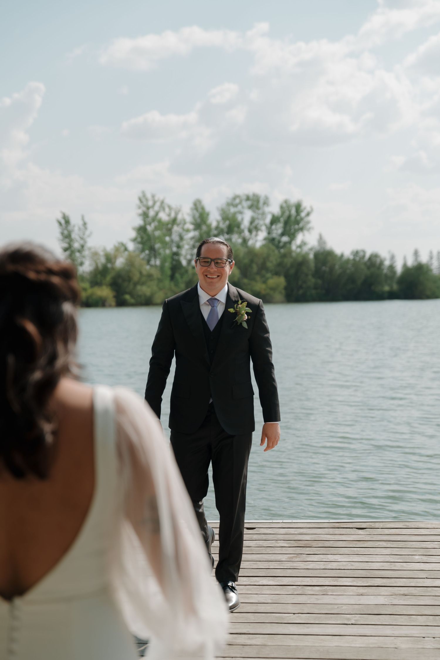 A groom in formal attire waits on a lakeside dock during an outdoor wedding ceremony.