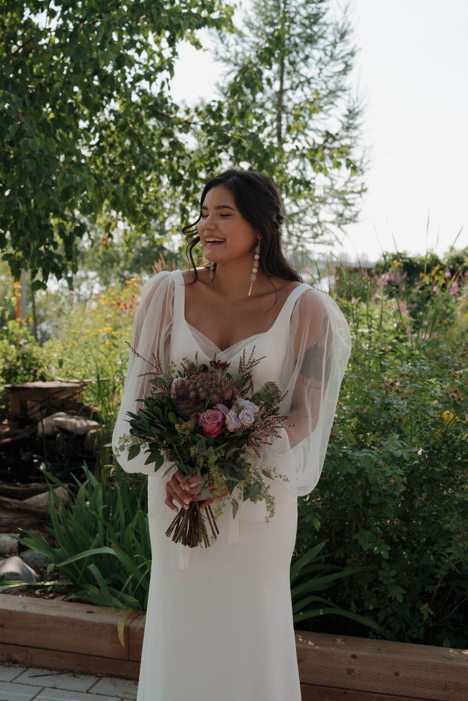 A joyful bride in a white dress holds a stunning bouquet of dark purple and pink flowers in a garden setting.