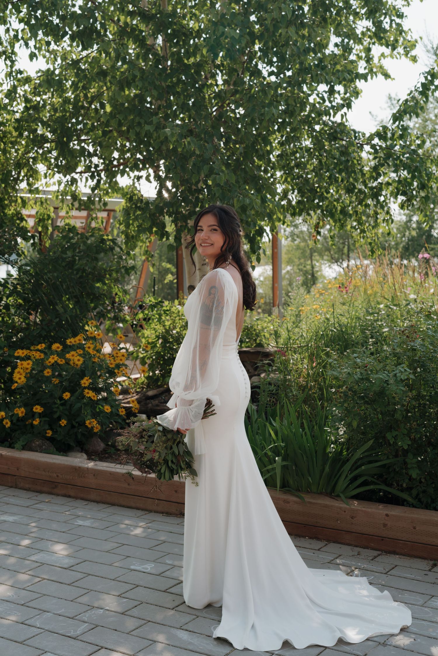 A person in a flowing white wedding gown poses in a garden setting with blooming marigolds and greenery.