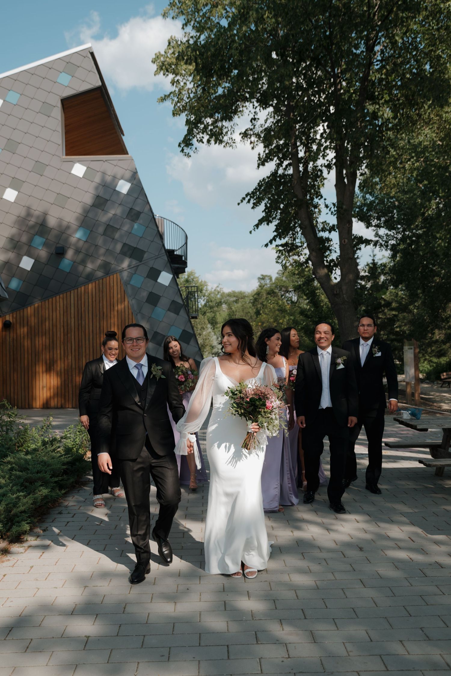 Wedding party walking along a modern paved path with a geometric building in the background.