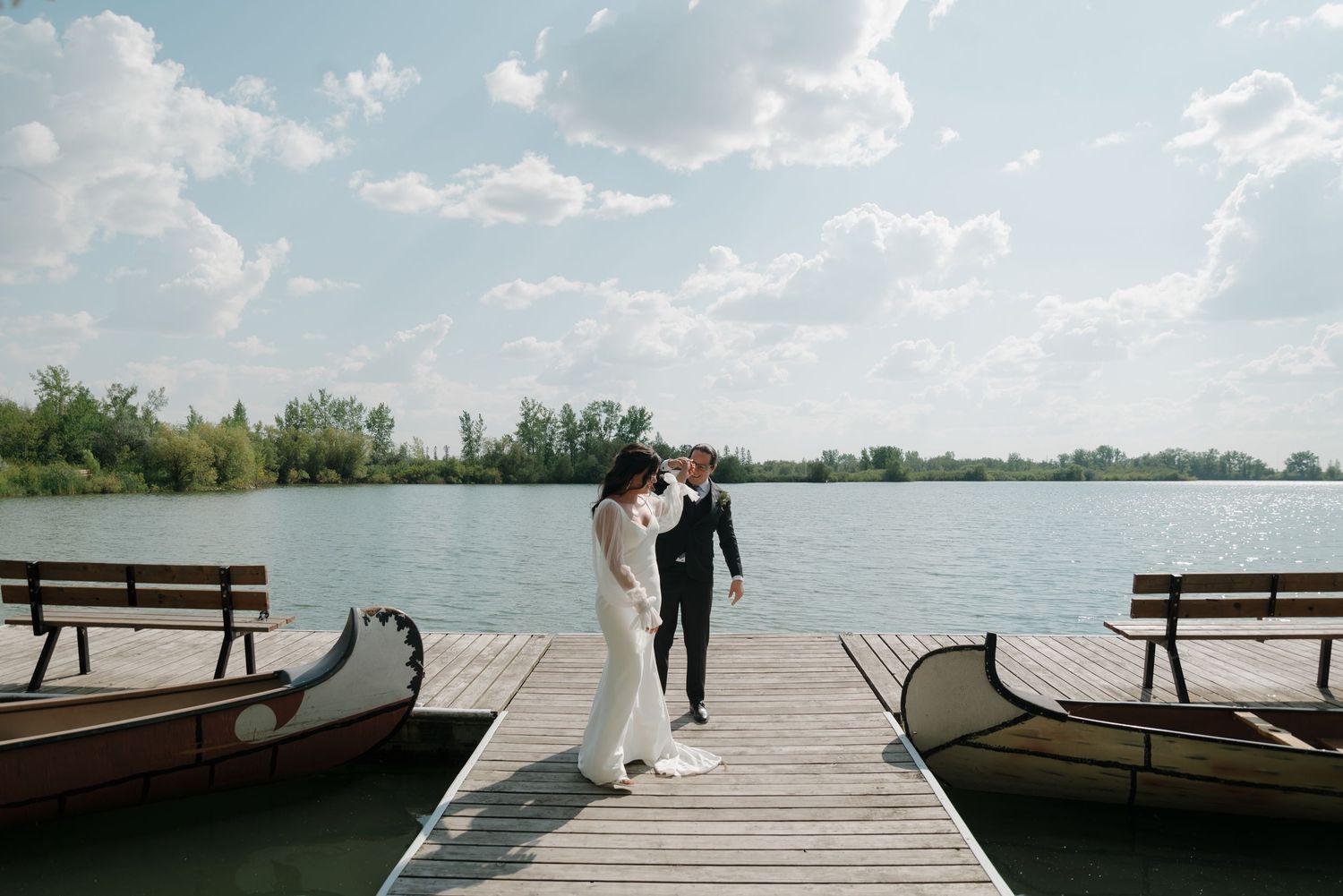 A wooden dock stretches out over calm waters under a bright blue sky with scattered clouds.