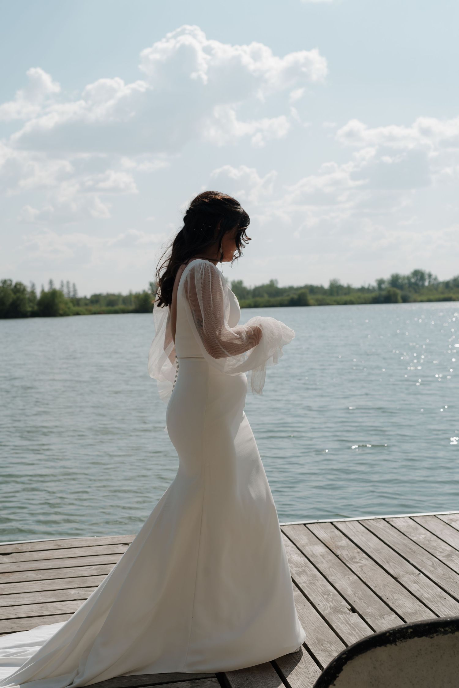 A flowing white wedding dress twirls gracefully on a wooden dock overlooking a serene lake.