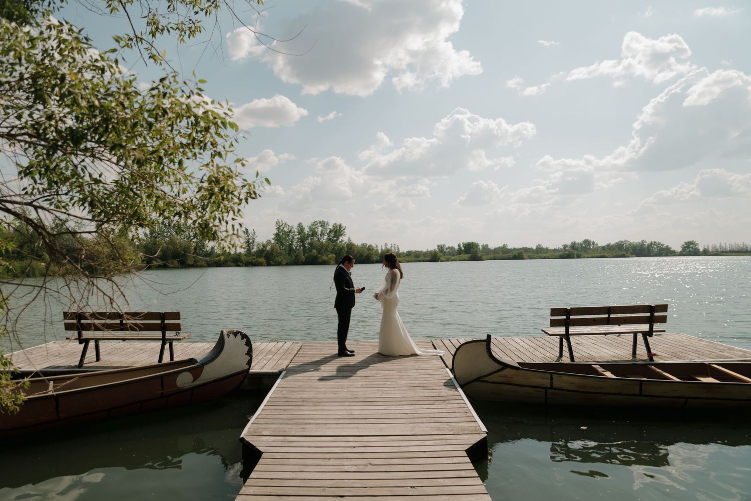 A wooden dock stretches out over a tranquil lake with benches and trees lining the shoreline on a sunny day.