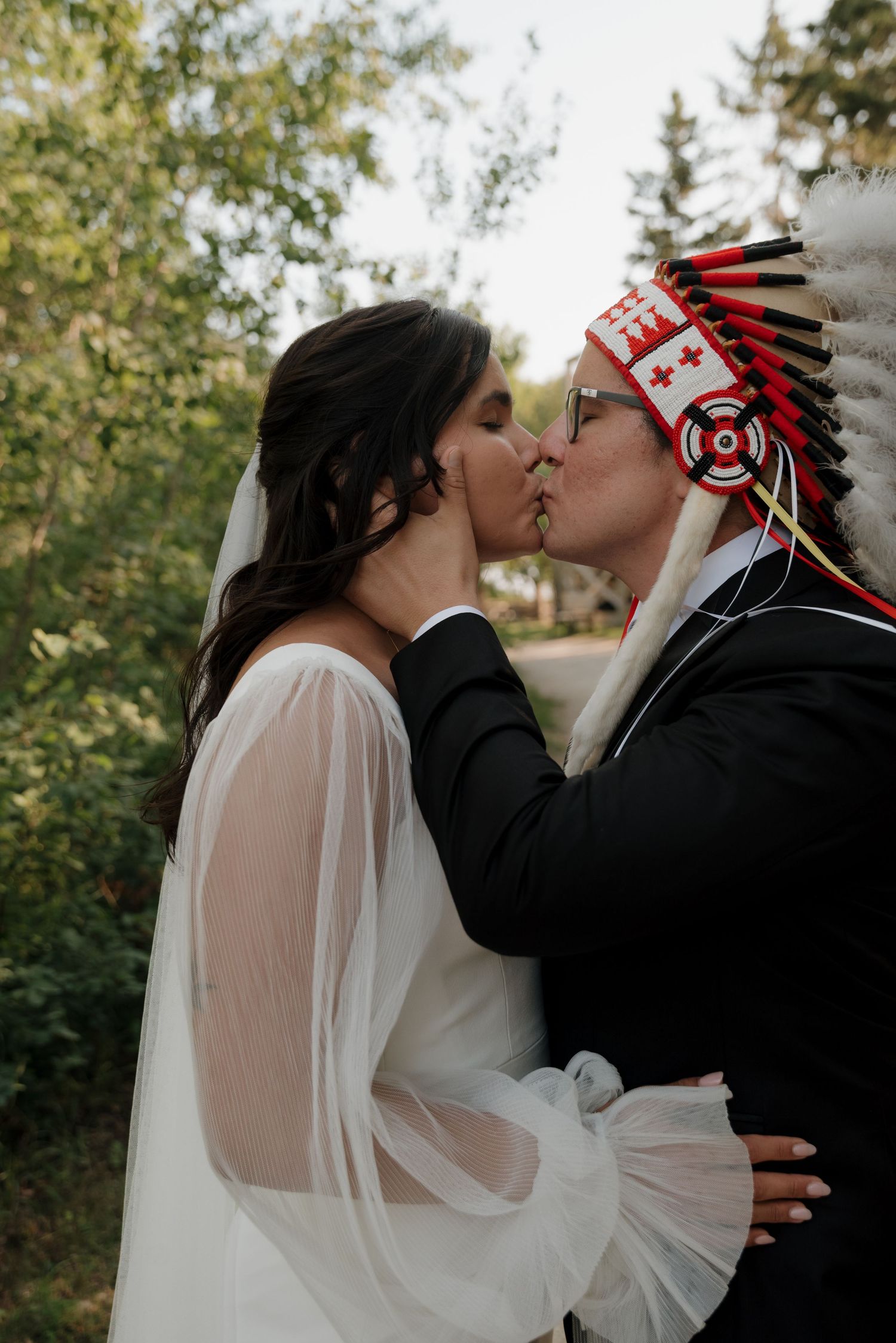 A couple shares a romantic kiss outdoors with one wearing a traditional headdress and the other in a flowing white veil.