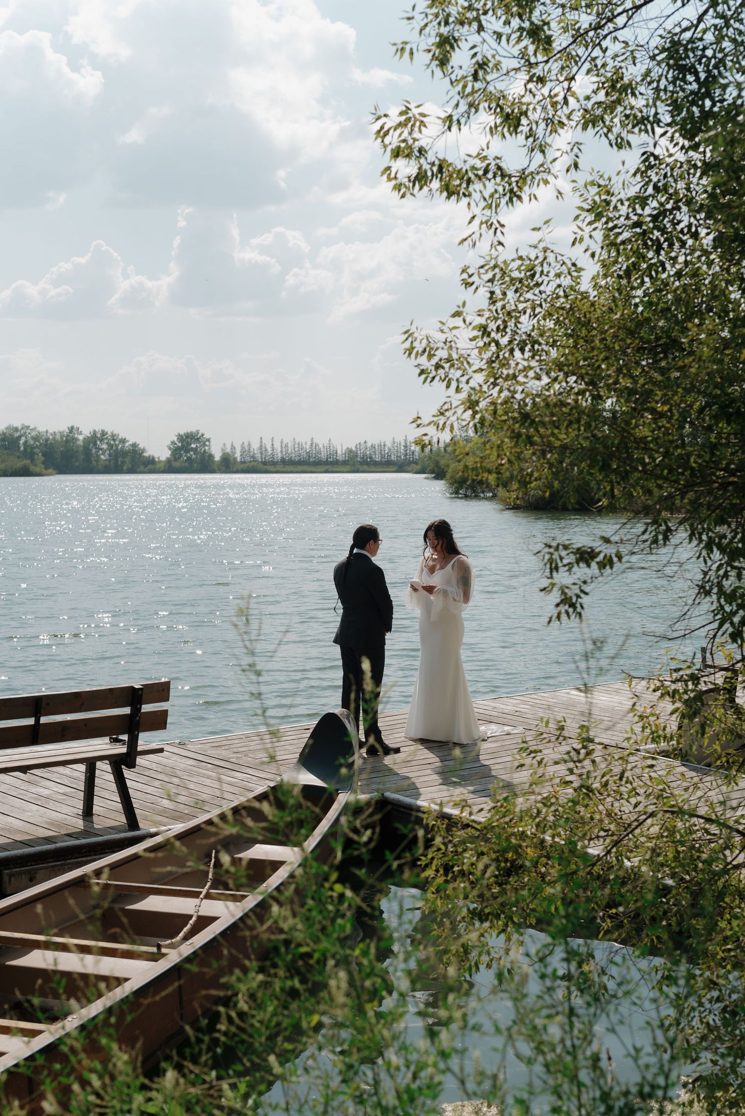 A couple shares a private moment on a lakeside dock surrounded by trees and sparkling water.