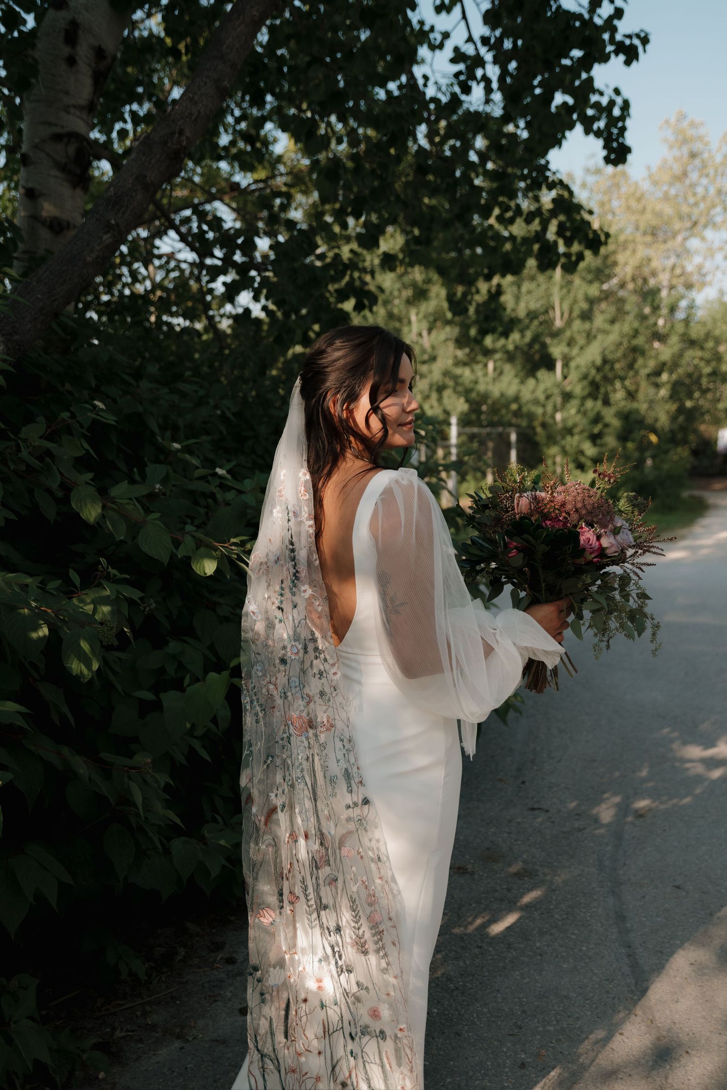 A bride in a flowing white dress with a delicate lace cape walks along a tree-lined path in natural lighting.