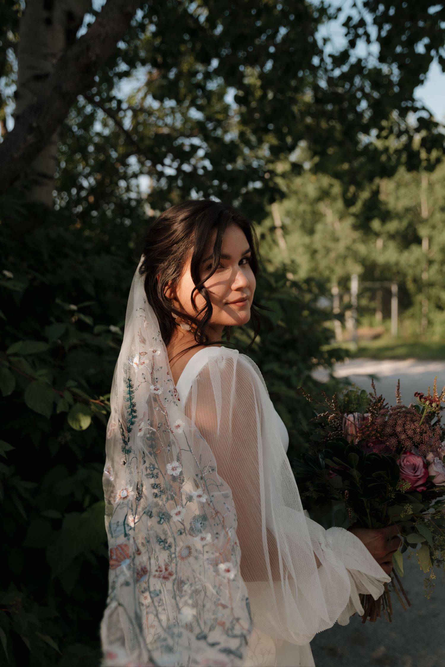 A bride in a flowing white veil with delicate floral embroidery poses in an outdoor garden setting at sunset.