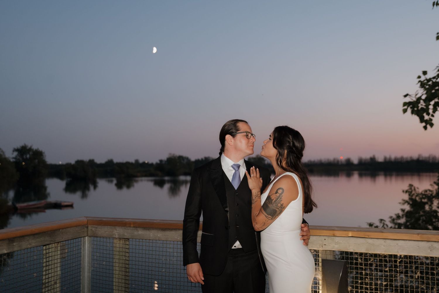 A couple embraces during sunset on a lakeside dock with tranquil water and sky in the background.