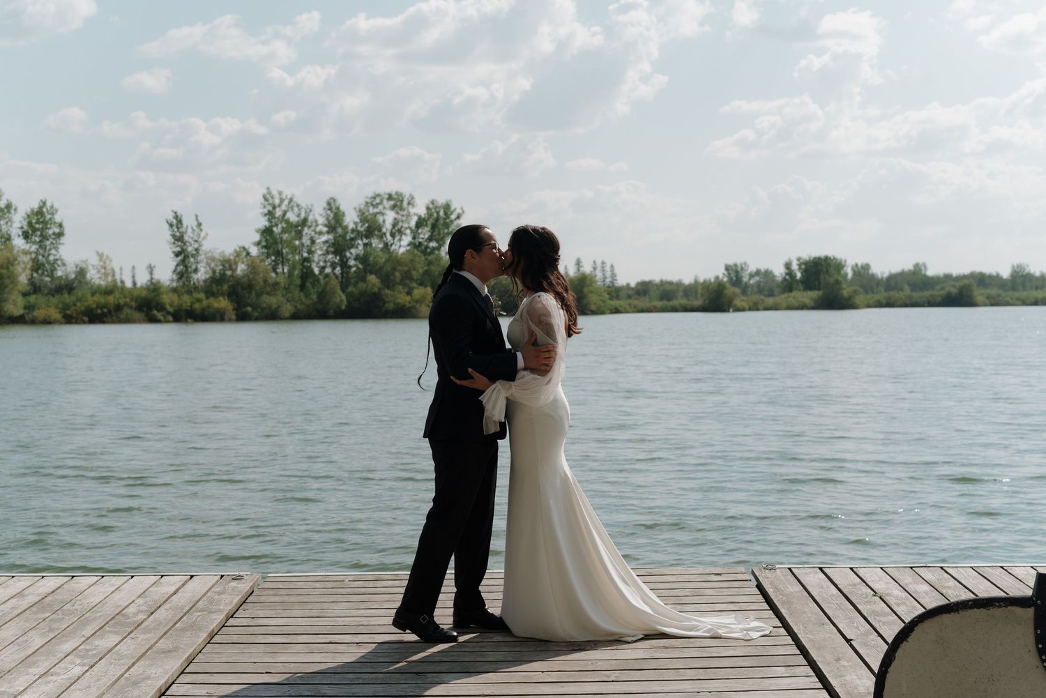 A wedding couple stands on a wooden dock overlooking a serene lake with trees in the background.