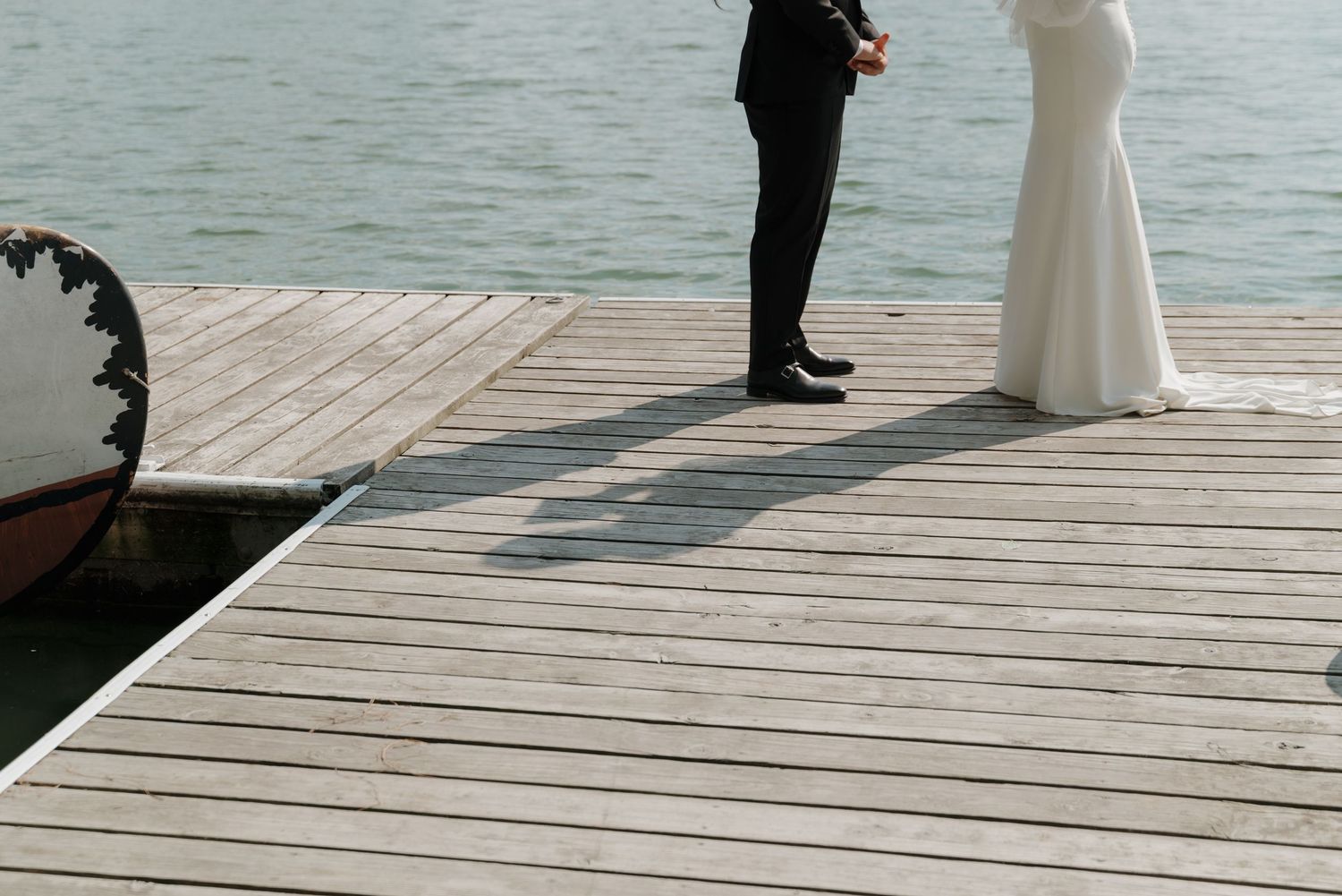 Two figures stand together on a wooden pier extending into calm waters on a bright day.