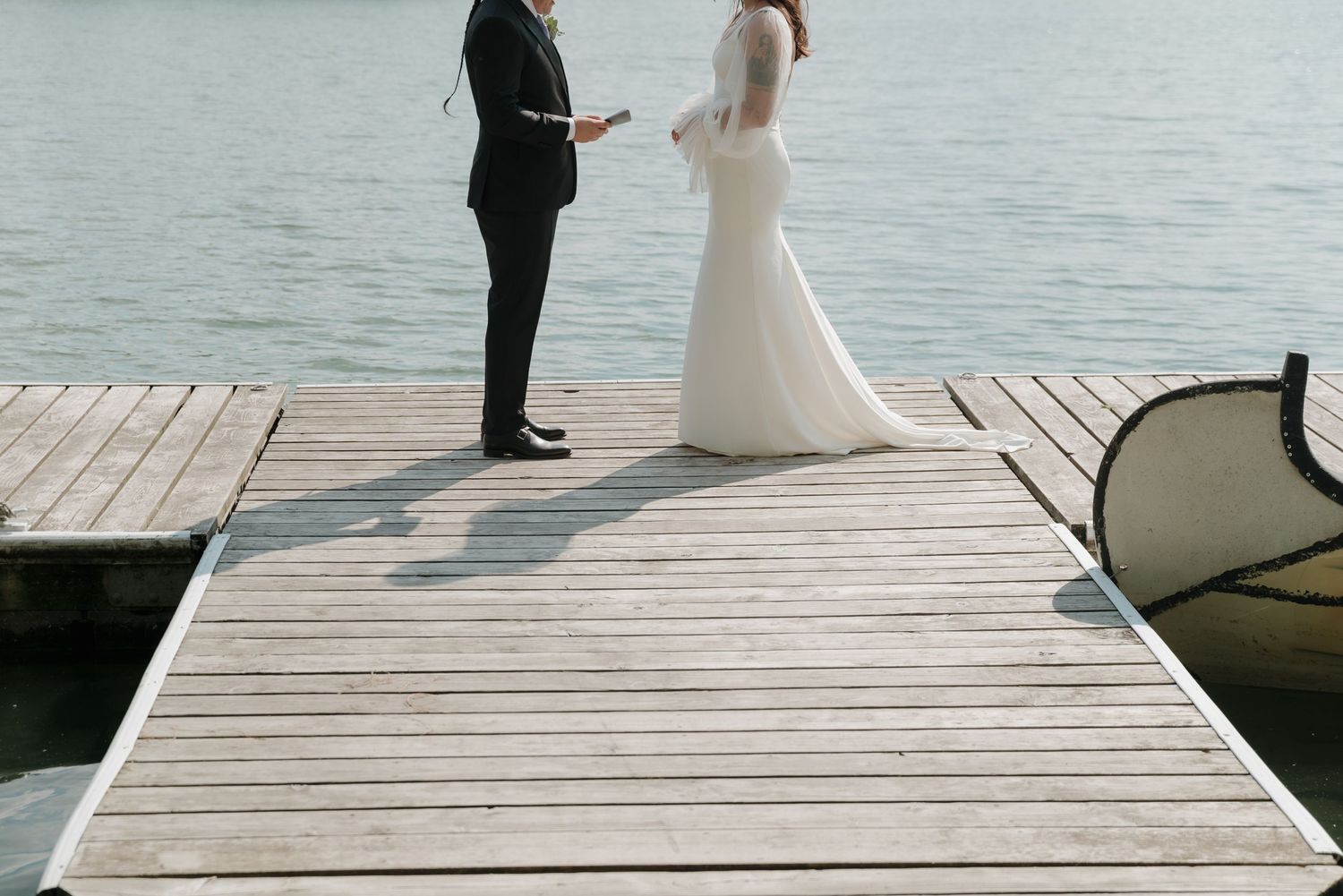 Two figures in formal attire stand together on a wooden pier overlooking a serene lake.