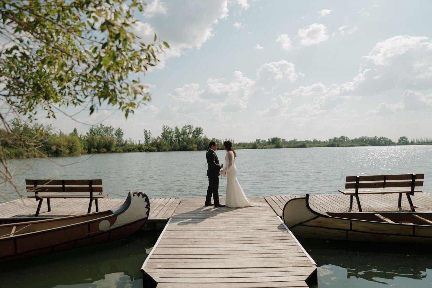 A wooden dock extends into a calm lake with benches and trees lining the waterfront on a sunny day.