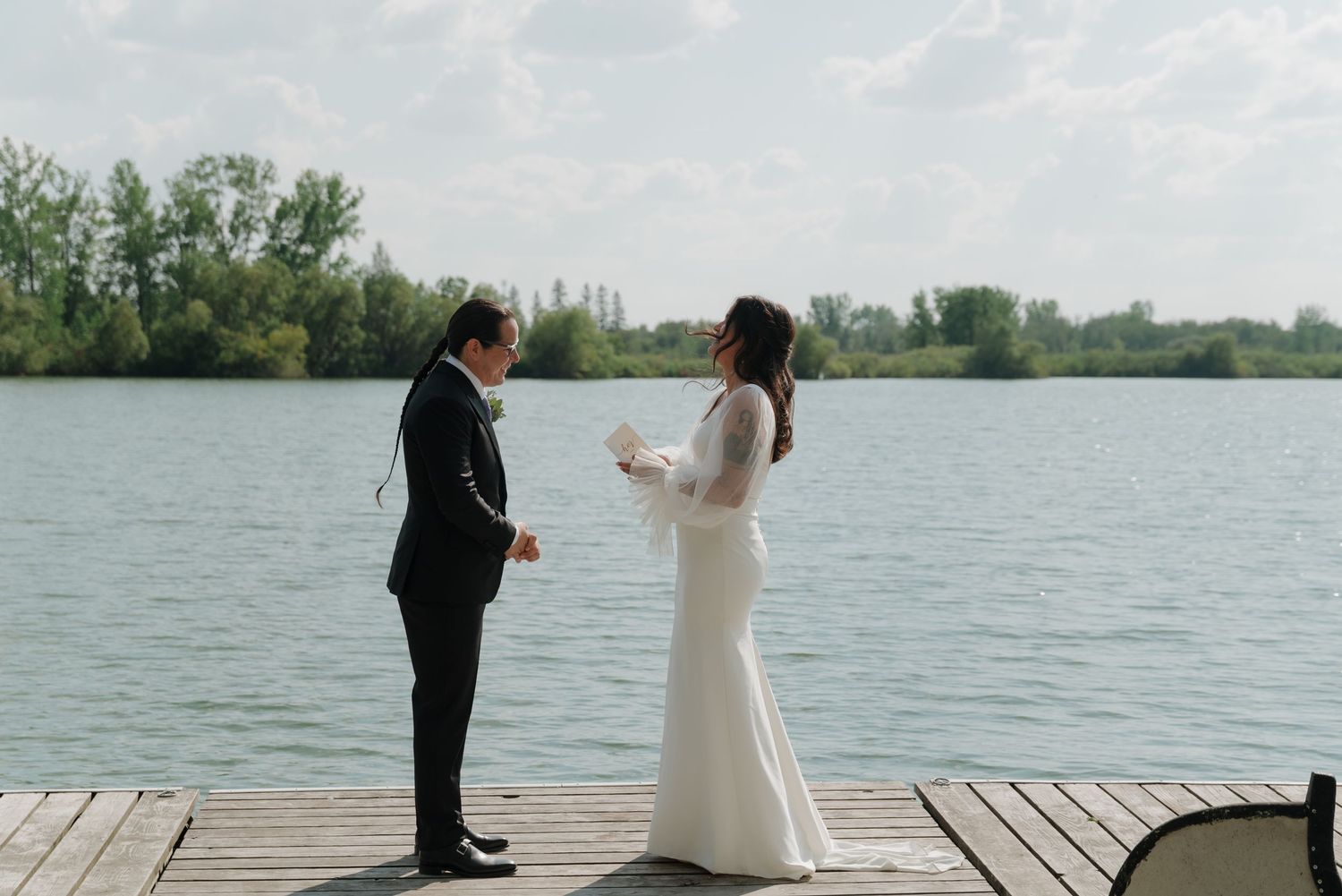 Couple standing on wooden dock by lake during outdoor wedding ceremony.