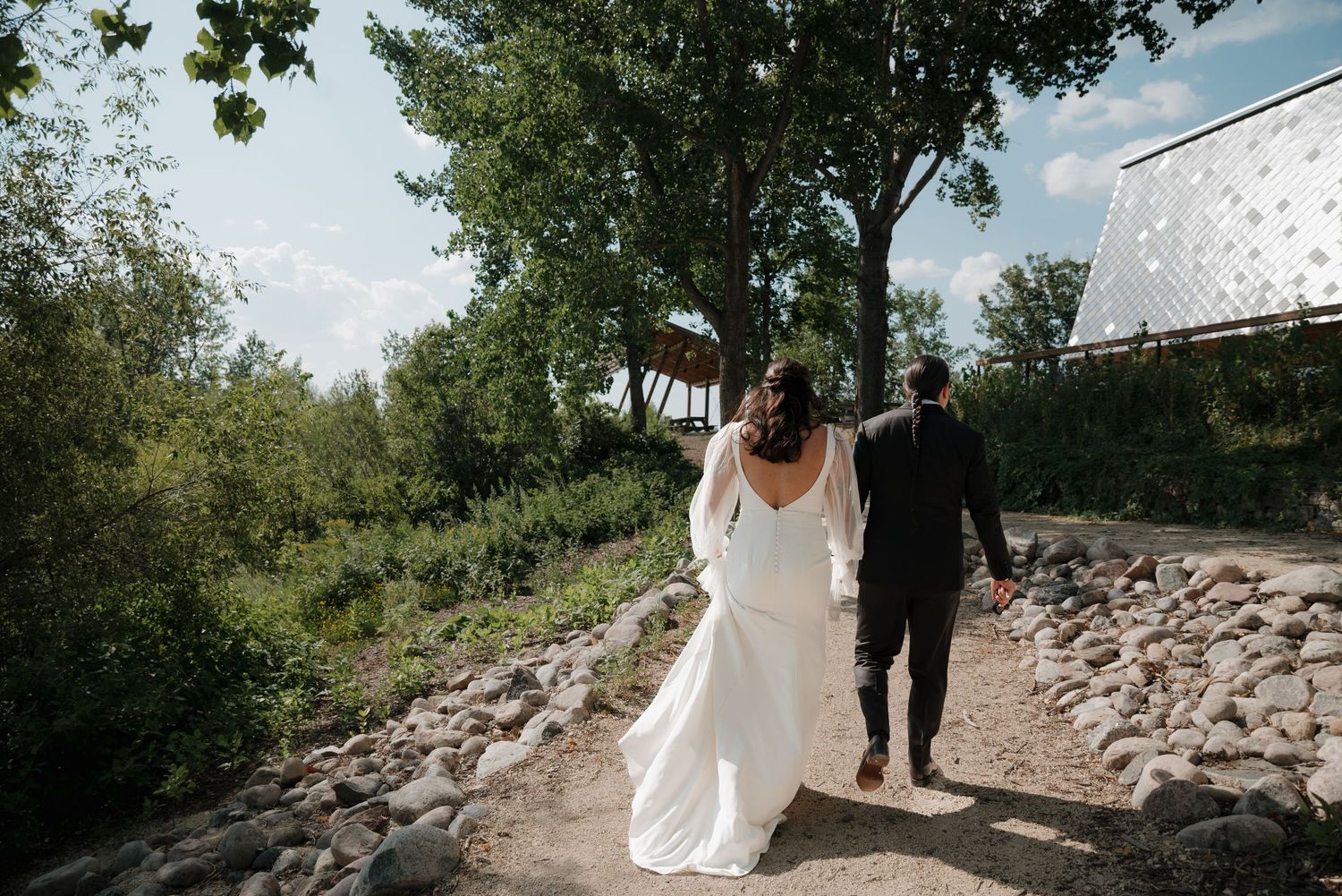Wedding party walks along a stone path outdoors with trees in the background on a sunny day.