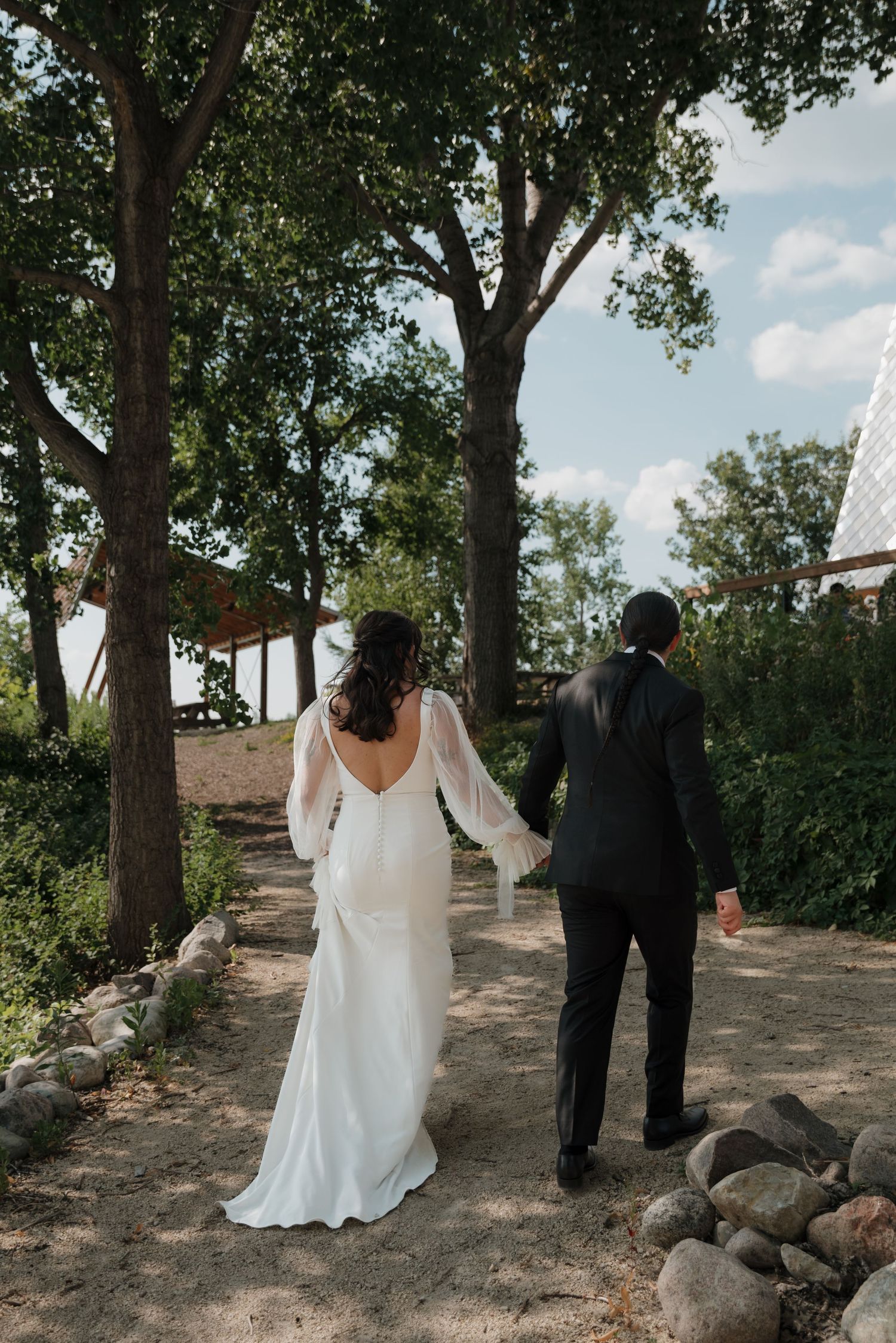 A couple in wedding attire walks along a tree-lined path on their special day.