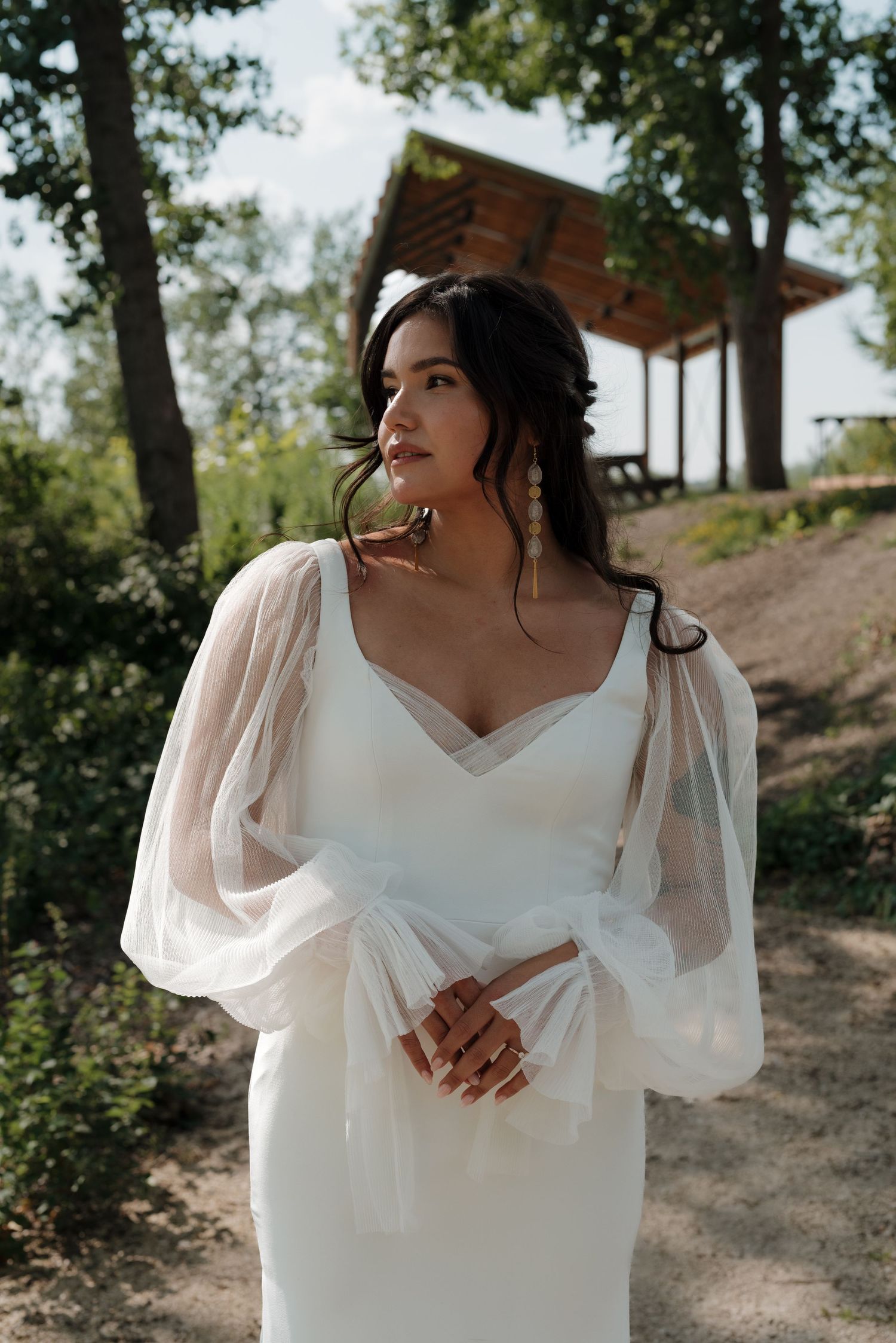 Bride in flowing white dress with sheer sleeves poses elegantly against natural outdoor backdrop.