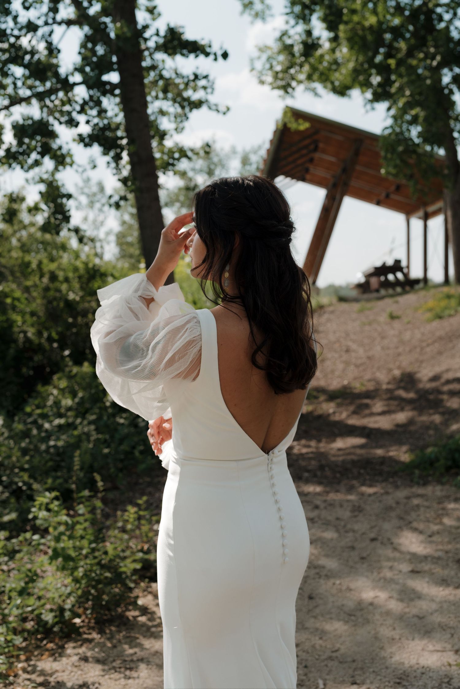 Bride in elegant white dress poses in a natural outdoor setting with trees and sunlight.
