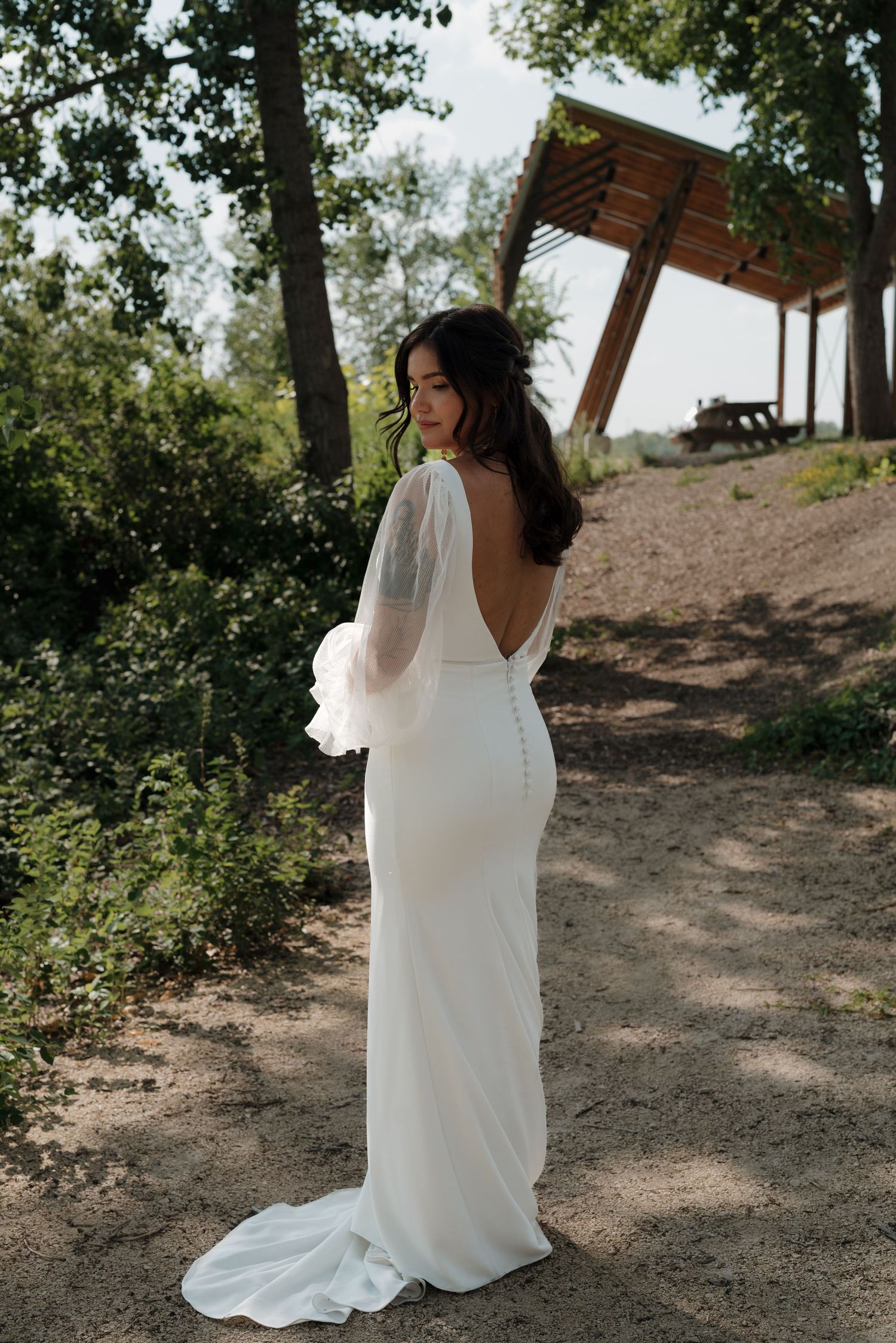 Bride in flowing white gown stands gracefully against a natural woodland backdrop.