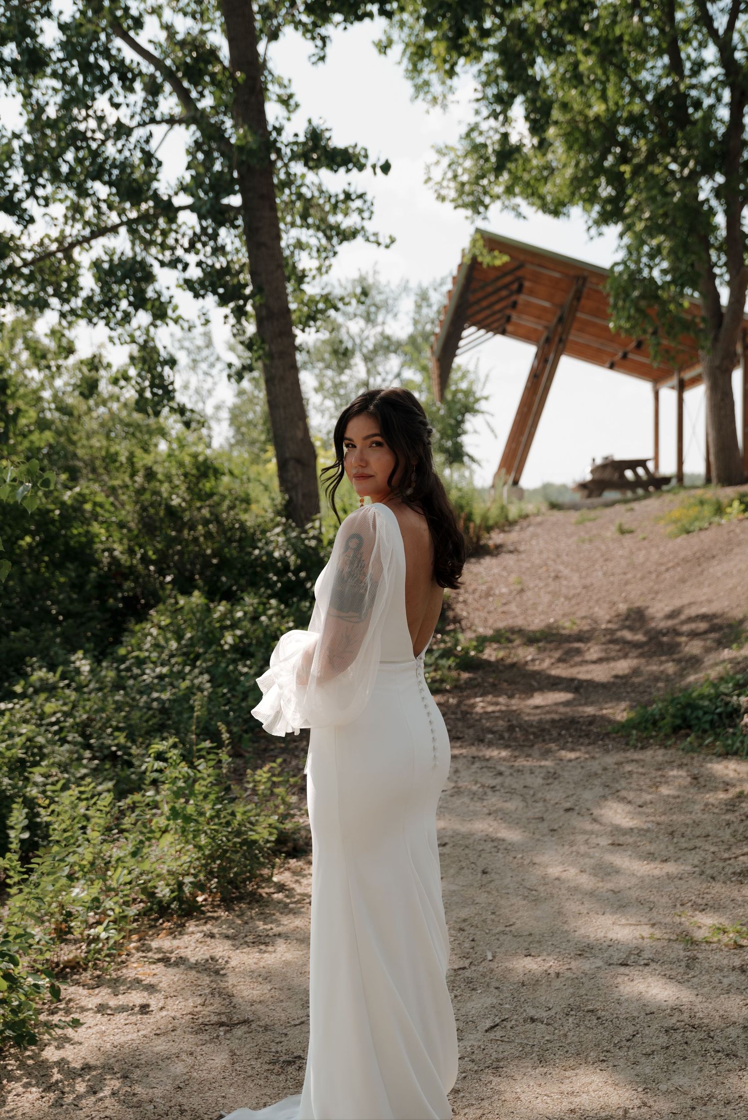 Bride in flowing white dress poses on hillside with trees in background.