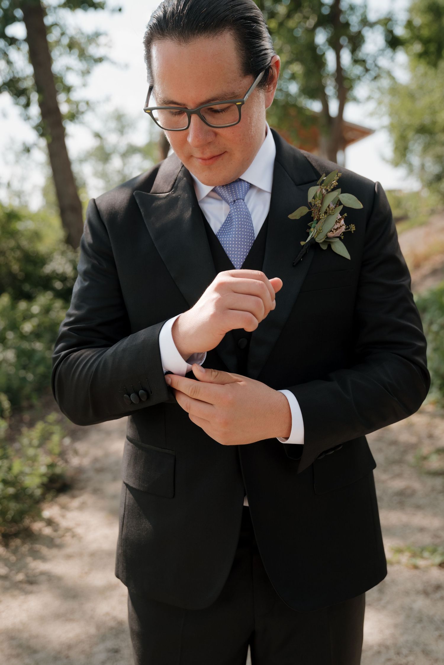 Groom in black suit and glasses adjusts his cufflinks during outdoor wedding photoshoot.