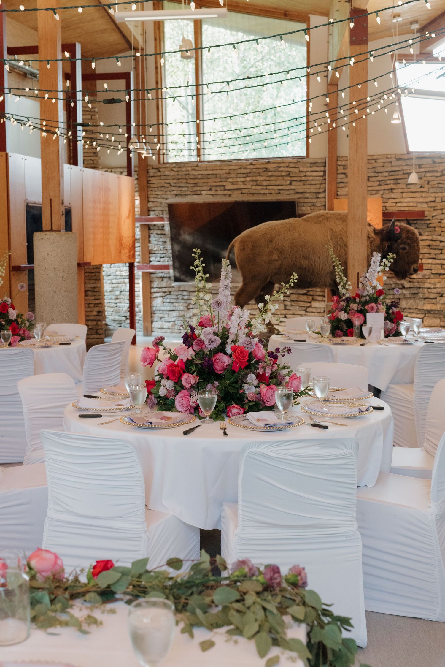 Wedding venue with exposed brick walls, string lights, and white-covered tables adorned with pink floral arrangements.