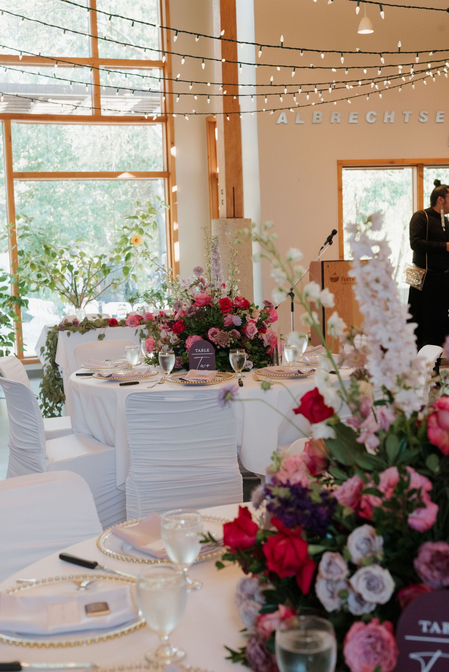 Wedding reception tables decorated with white linens and vibrant pink and purple flower arrangements under twinkle lights.