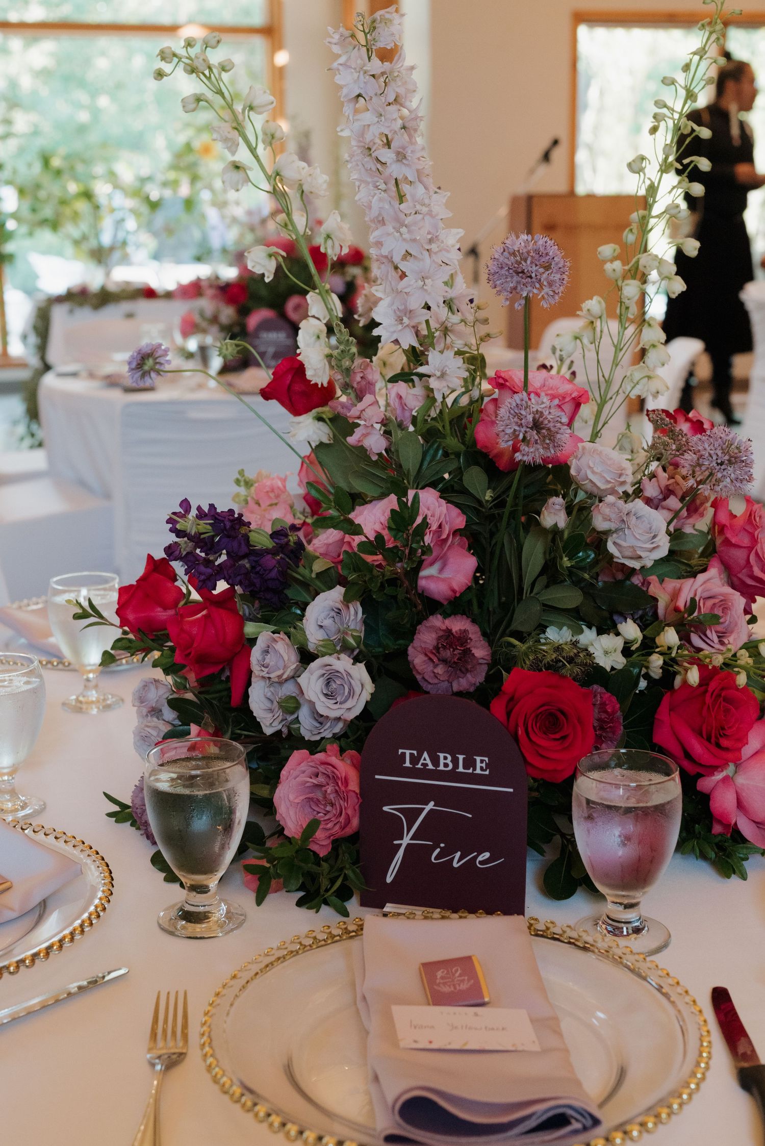 Elegant wedding table setting with pink and burgundy floral centerpieces, gold-rimmed plates, and burgundy table numbers.