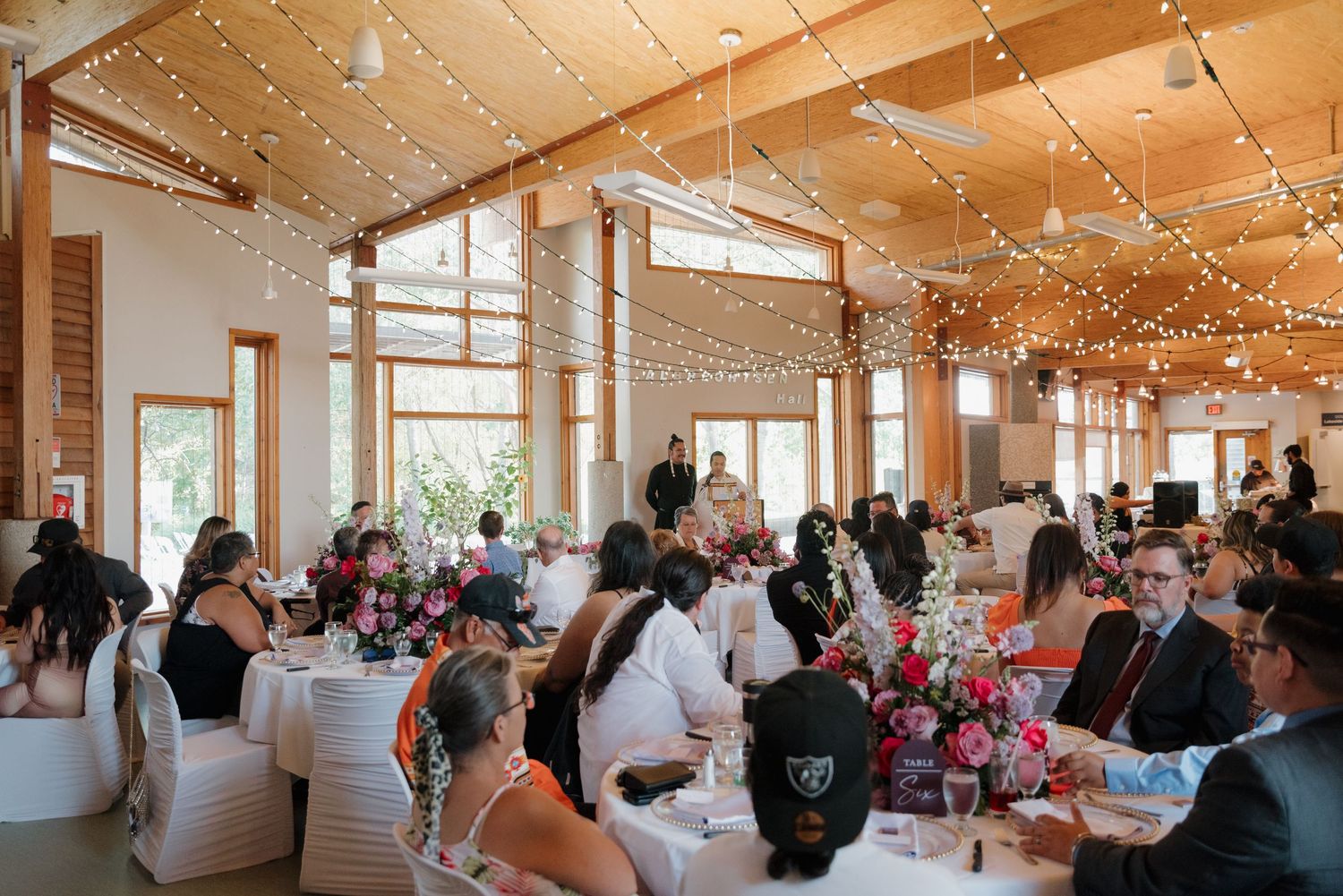 Wedding reception guests seated at tables in a bright modern venue with string lights and high ceilings.