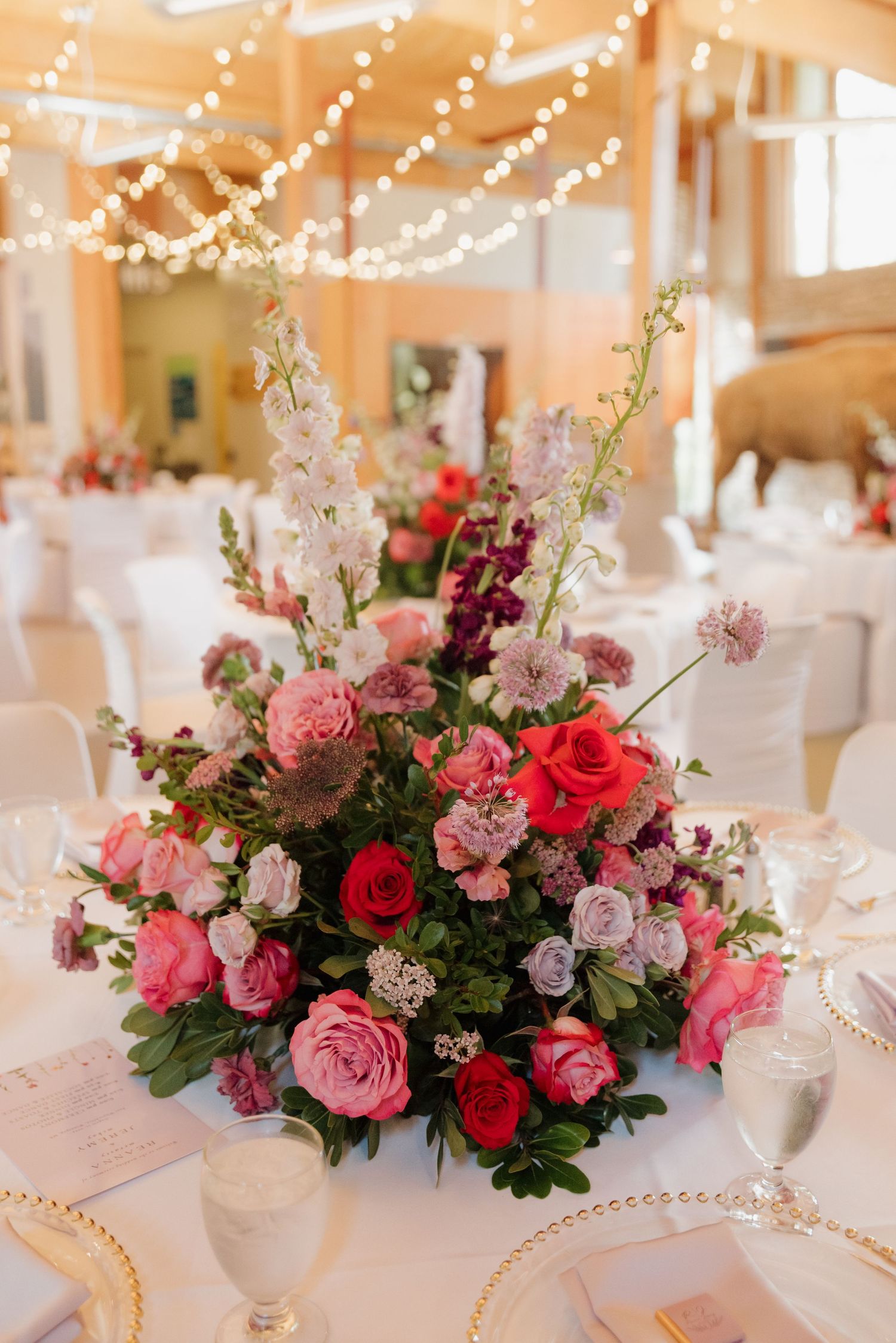Elegant floral centerpieces with pink and red roses adorn white-clothed tables at a wedding reception.