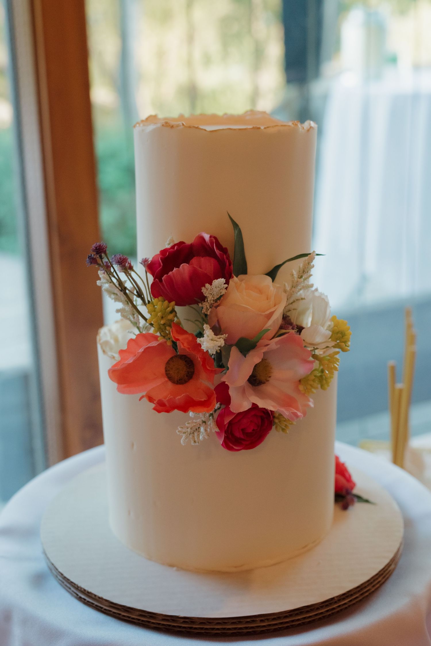 Elegant white wedding cake decorated with fresh red and pink flowers on a simple cake stand.
