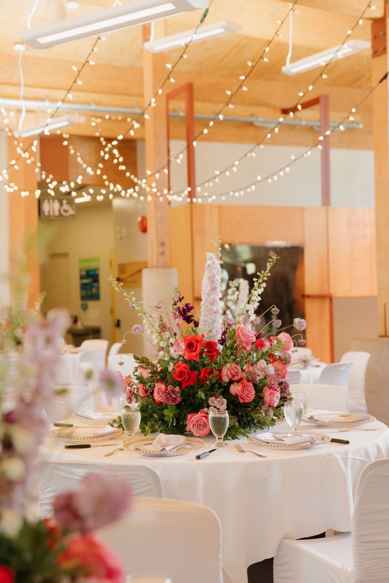 Wedding reception venue decorated with string lights and pink floral arrangements on round tables with white linens.