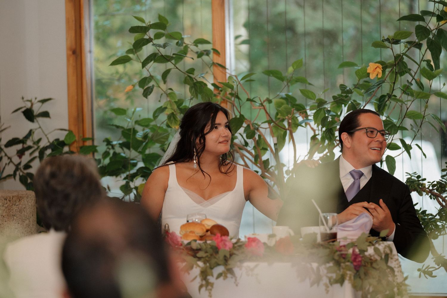 Wedding reception in a bright greenhouse-style venue with lush plants and wooden beams.