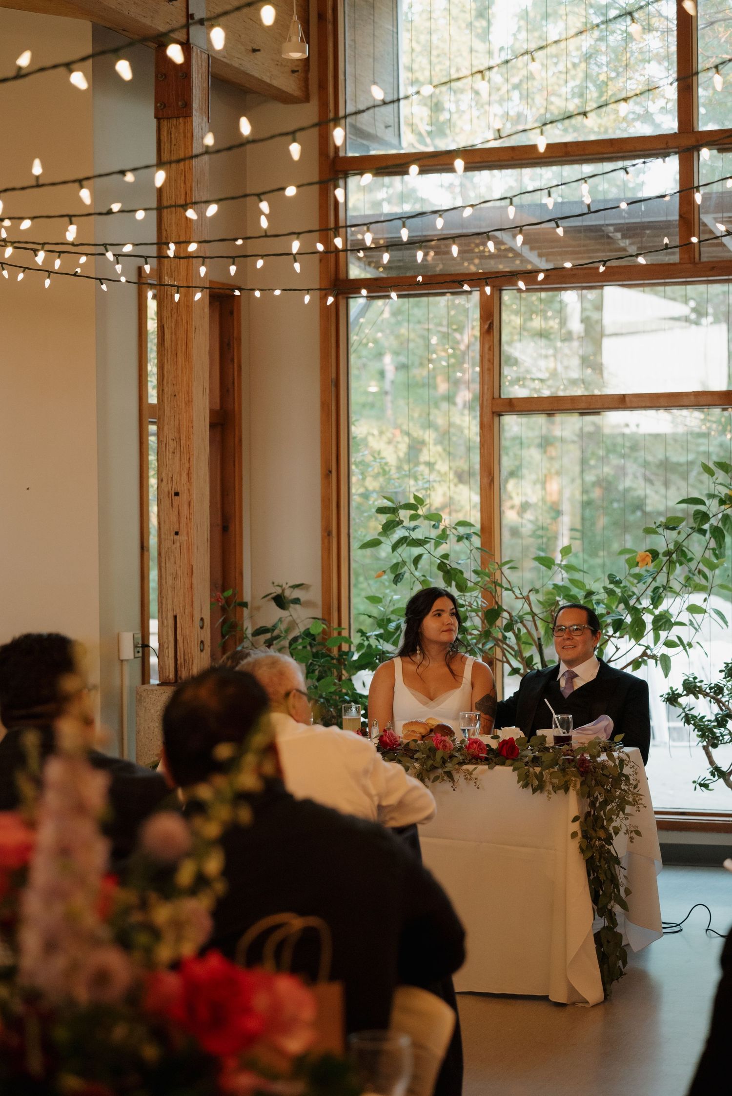 Wedding reception venue with string lights and natural lighting streaming through large windows framed by wooden beams.