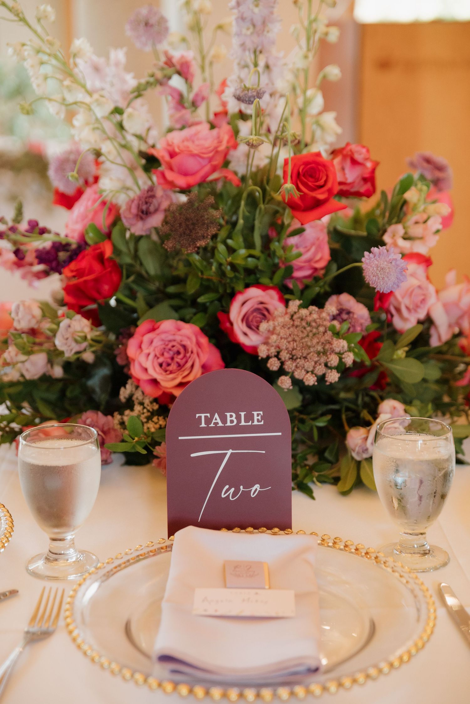 Elegant wedding table setting with pink roses, purple table numbers, and gold charger plates on white linens.
