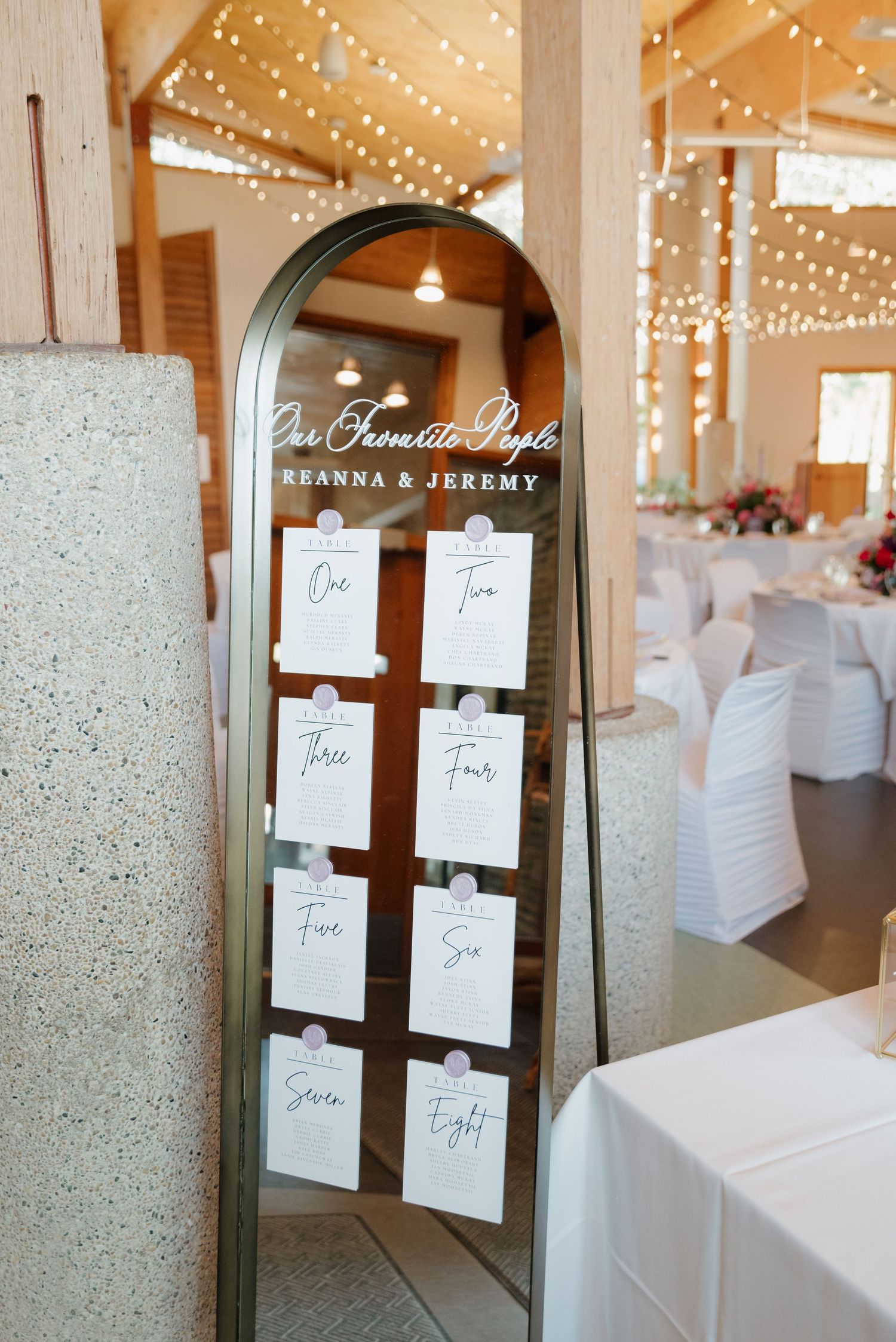 Wedding seating chart displayed on a rustic mirror with twinkle lights in the background.