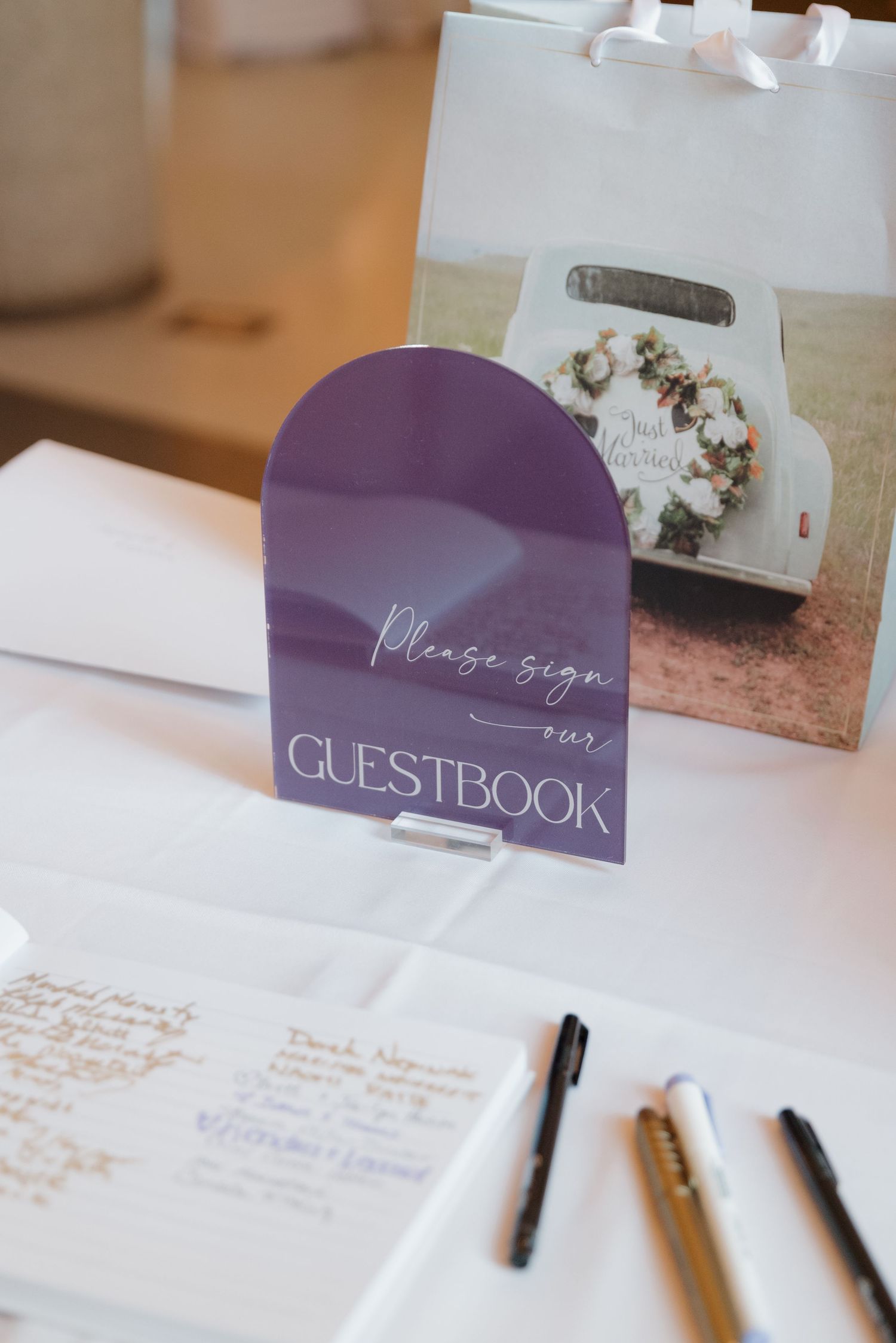 Purple and white wedding guestbook station with pens and decorative signage.