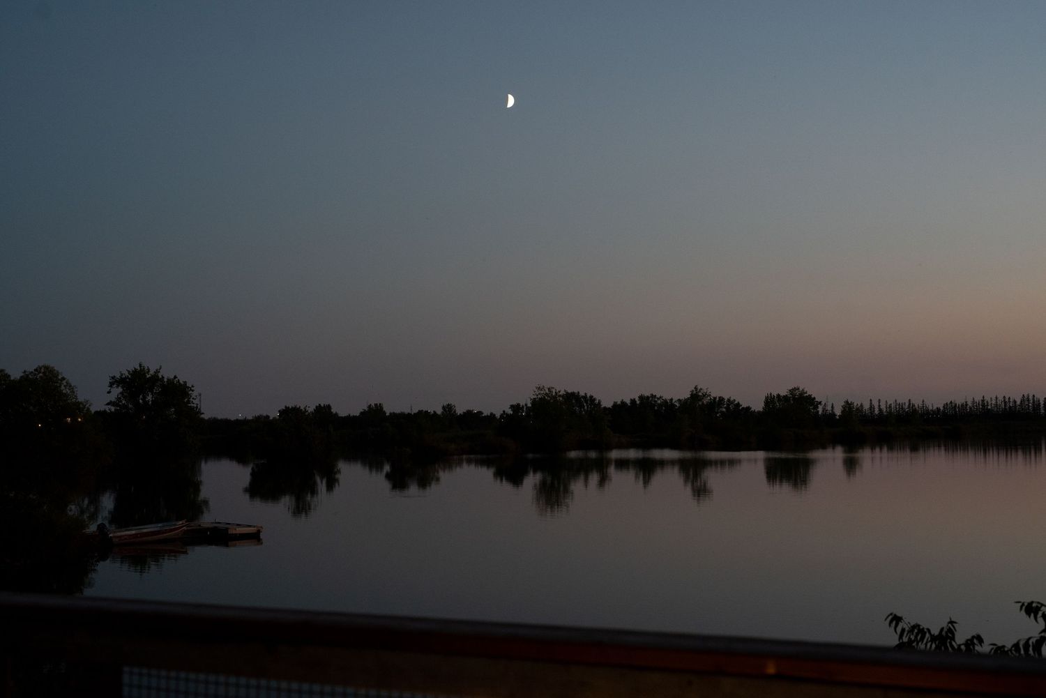 A serene panoramic view of a lake at sunset with silhouettes of trees reflected in the calm water.