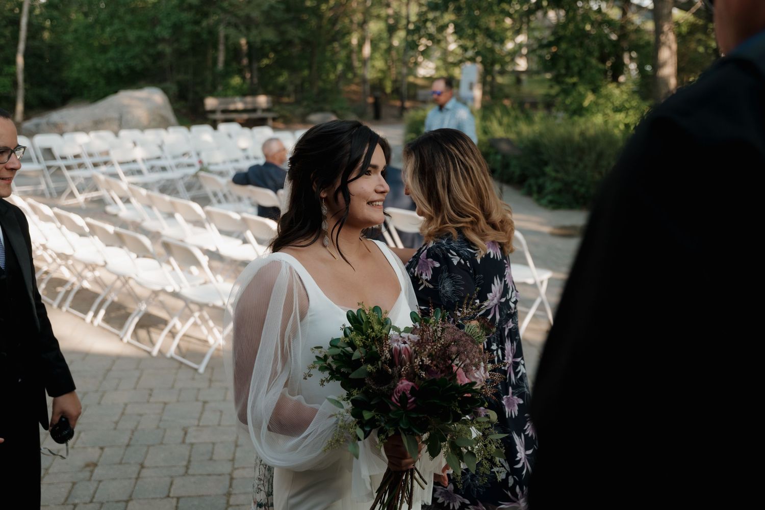 A wedding ceremony in progress with guests seated in white chairs on a stone patio surrounded by greenery.