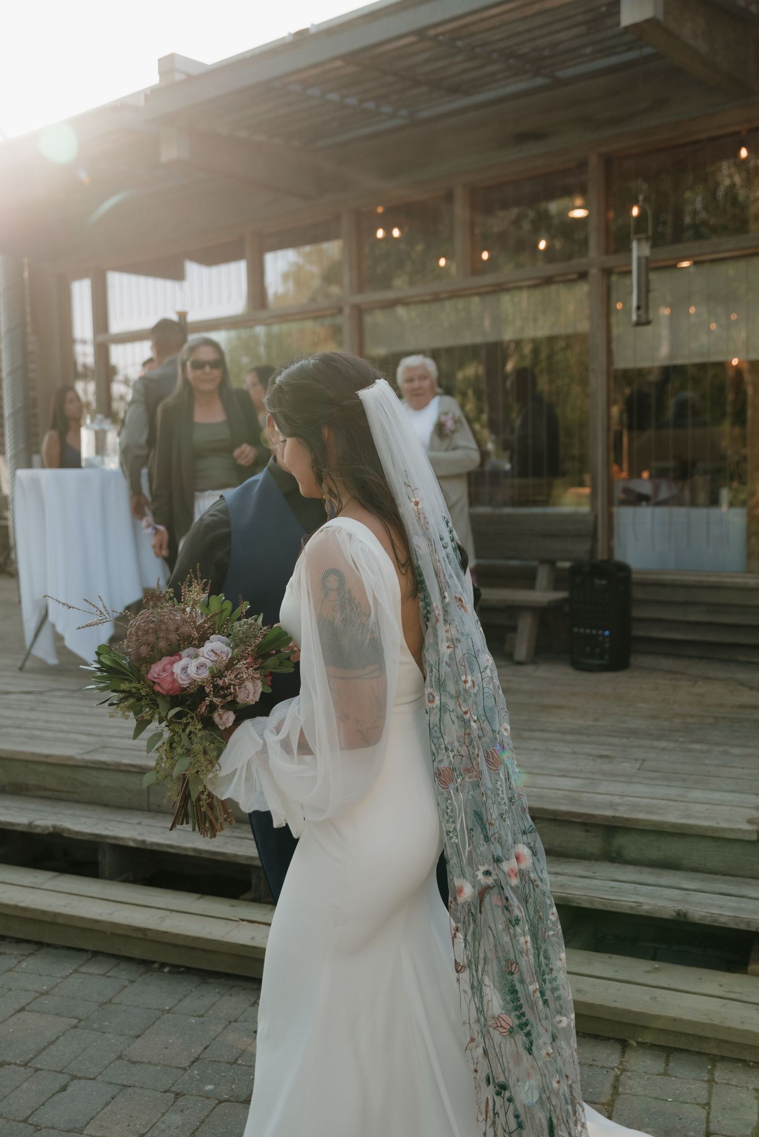 Wedding party posing outdoors on wooden boardwalk during sunset with elegant attire and floral arrangements.