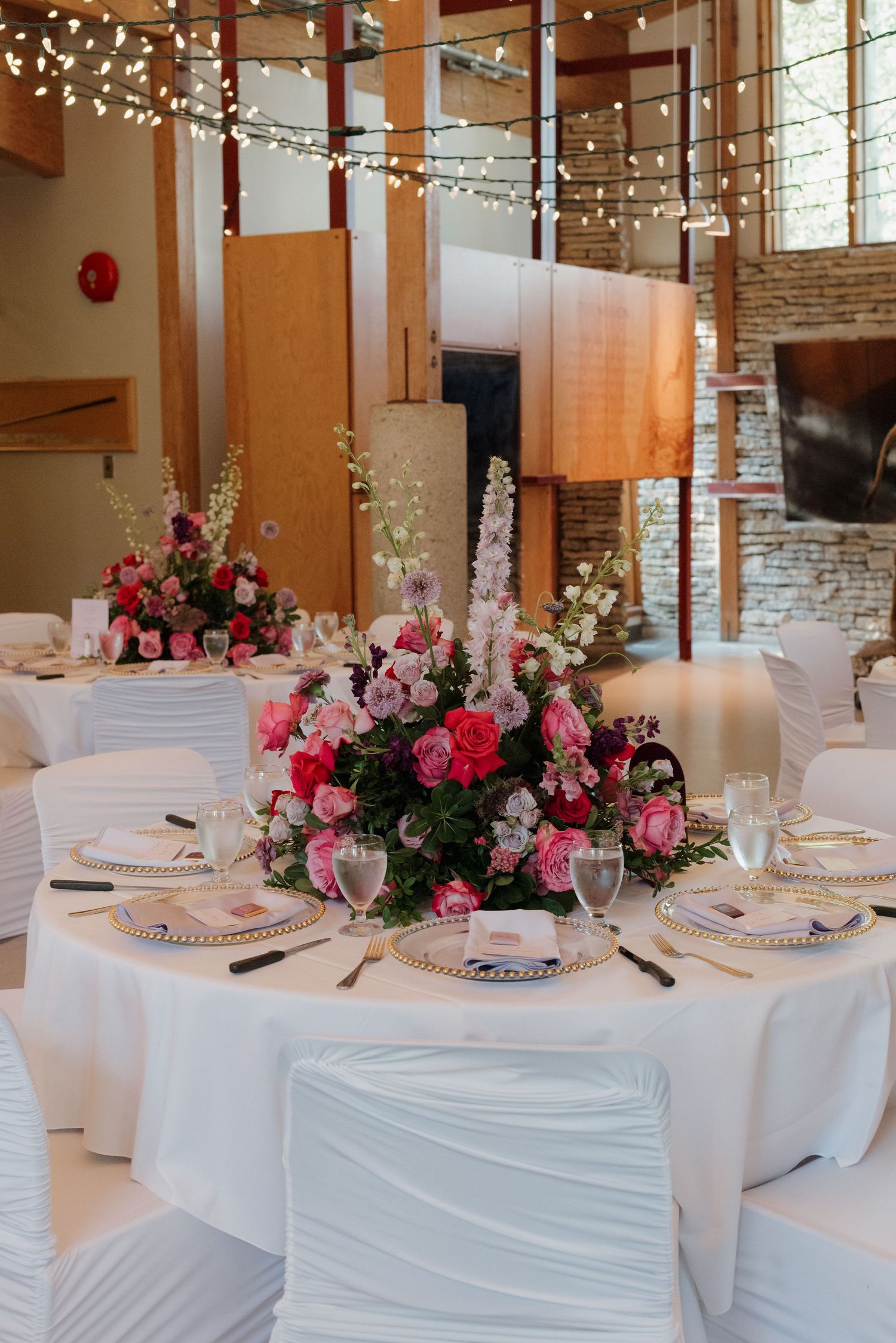 Elegant wedding reception tables with white linens and pink floral centerpieces under string lights in rustic venue.