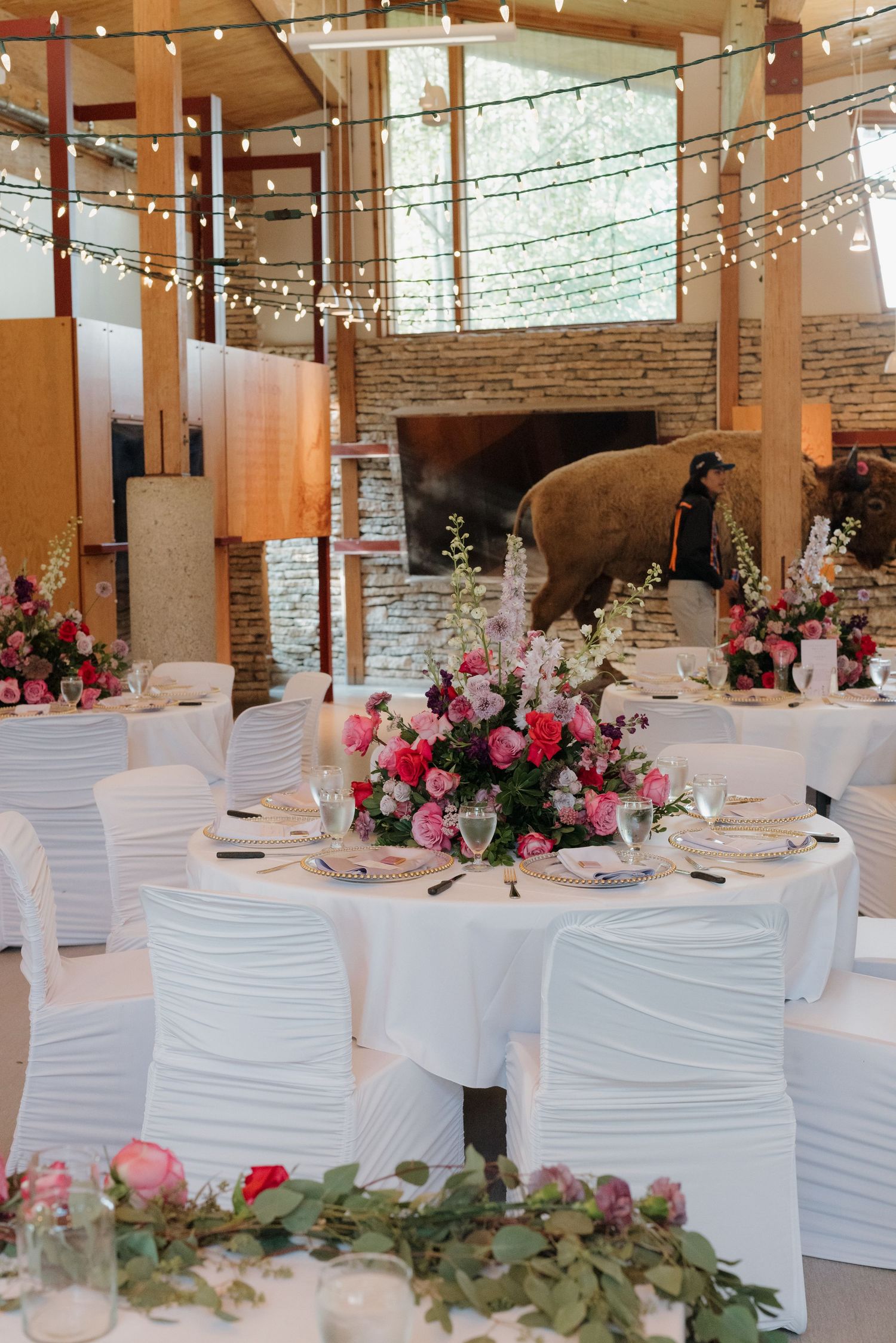 Wedding reception setup with round tables, white chair covers and pink rose centerpieces in industrial-style space.